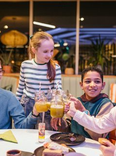 Four children clink their glasses together at a table; they are surrounded by sweet treats and balloons on the table.