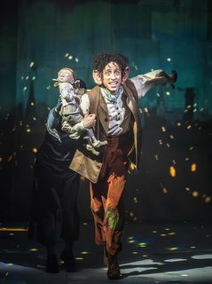 A man with dark brown curly hair and big ears wearing a white shirt and orange/brown trousers with a white shirt and brown waistcoat holding a small puppet on his arm in a running pose