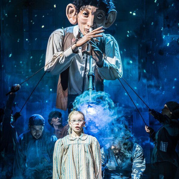 A giant puppet with short dark curly hair wearing a white shirt with a brown waistcoat on holding a big pipe to his lips. In front of the puppet there is a girl with stripped pyjamas on with a shocked expression on her face