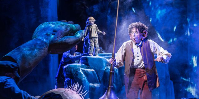 A big puppet with spiky hair on the ground holding up his big arm towards a man with short curly hair wearing a white shirt, waistcoat and orange/brown trousers looking quite shocked holding up a long pipe. In the background a small puppet is standing on a large rock