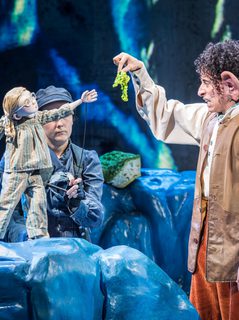 A man with dark curly hair and big ears wearing a white shirt, brown waistcoat and orange trousers holding up a green piece of fabric in front of a small girl puppet wearing striped pyjamas. The puppet is being operated by two women wearing dark clothes and a hat