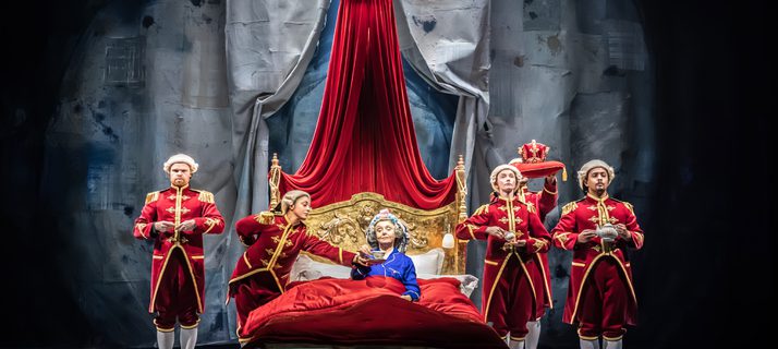 A woman with grey hair in purple pyjamas lying in a bed with a red duvet and gold-plated headrest holding a cup of tea. Next to her are five people in servant uniform