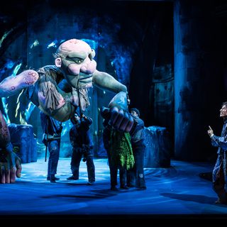 A man with curly black hair wearing a long cloak and a backpack pointing his finger up to a giant green and pink puppet which is being operated by people wearing dark clothes