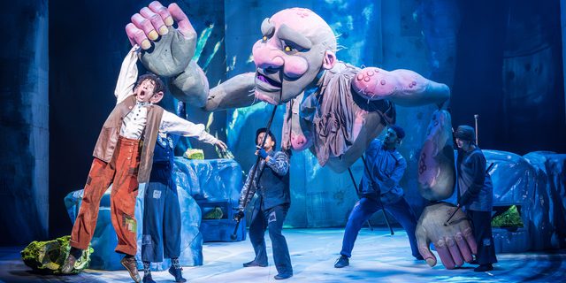 A man with curly black hair wearing orange/brown trousers and a white shirt and brown long waistcoat being held up in the air by a giant green and pink puppet. Four people in dark clothes and hats are stood below operating the puppet