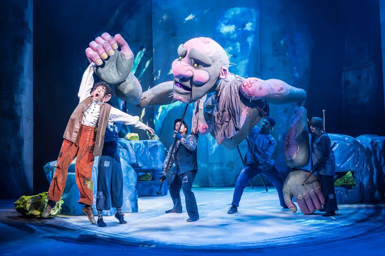 A man with curly black hair wearing orange/brown trousers and a white shirt and brown long waistcoat being held up in the air by a giant green and pink puppet. Four people in dark clothes and hats are stood below operating the puppet