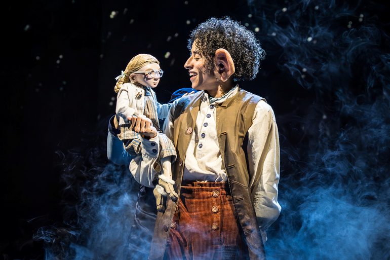 A man with short curly dark hair and big ears wearing a white shirt, orange/brown trousers and waistcoat, holding a small puppet of a girl