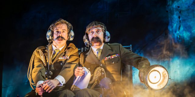 Two men sat next to each other wearing military uniform, both with dark short hair and moustaches and with shocked expressions on their faces. One of the men has a considerably larger moustache and is holding a map and large torch