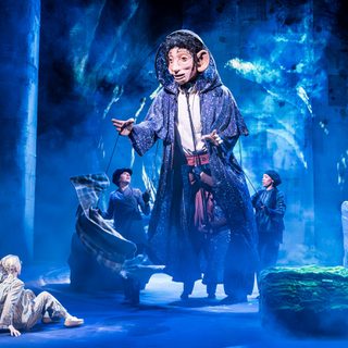 A small girl with blonde hair wearing stripped pyjamas is sat down on the ground looking at a giant man puppet that is wearing a long purple cloak being operated by two women in dark clothes and hats