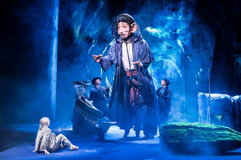 A small girl with blonde hair wearing stripped pyjamas is sat down on the ground looking at a giant man puppet that is wearing a long purple cloak being operated by two women in dark clothes and hats