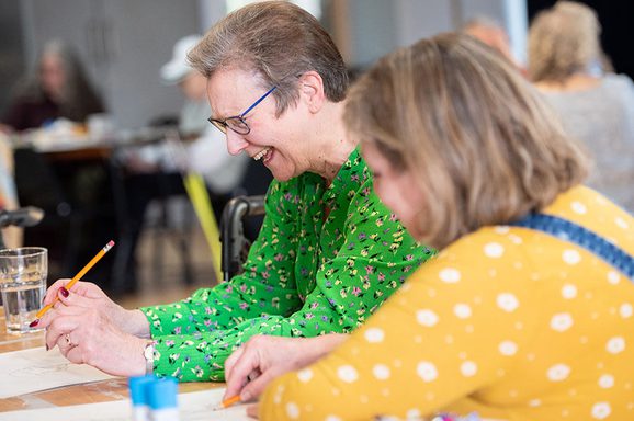 Two older women sit at a desk writing and laughing together.