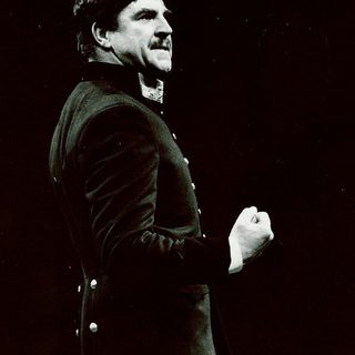 An actor stands side with a clenched fist, wearing a hat and military uniform