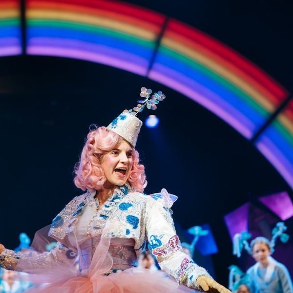 Production photo from The Wizard of Oz. A girl with pink curly hair wearing a silver and blue embellished jacket and matching hat holds a long, gold, sparkling wand. She looks off to the right with her mouth open, like she is singing. In the background, there is a rainbow in lights. Other young performers are out of focus, looking at her in awe.