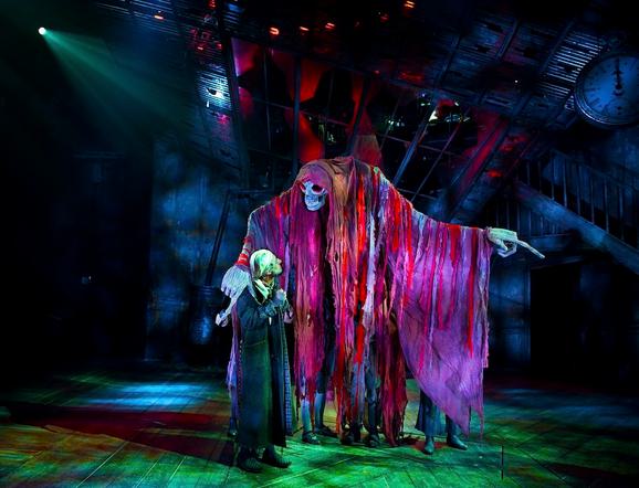 Production photo from A Christmas Carol. A huge skeleton puppet looms over the stage, draped with a huge cloak of red and purple rags. Below stands a man in a long, dark coat, slippers and a white nightcap, looking up at the puppet.