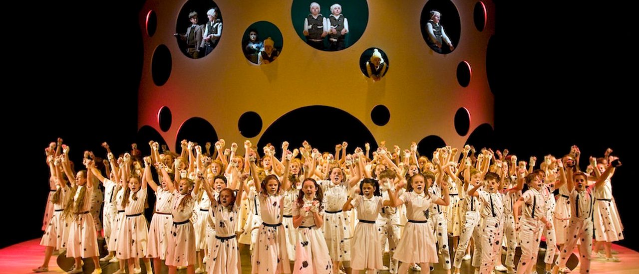 Production photo from The Hundred and One Dalmatians. The stage is full of young performers dressed as Dalmatians with their arms in the air, singing.
