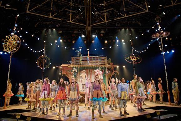 Production photo from Pinocchio. A group of young performers wearing colourful costumes all stand proudly around the stage, looking out to the audience and singing.