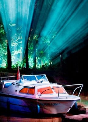 A white boat sits at a riverbank. Light streams in blue and white through trees behind it.