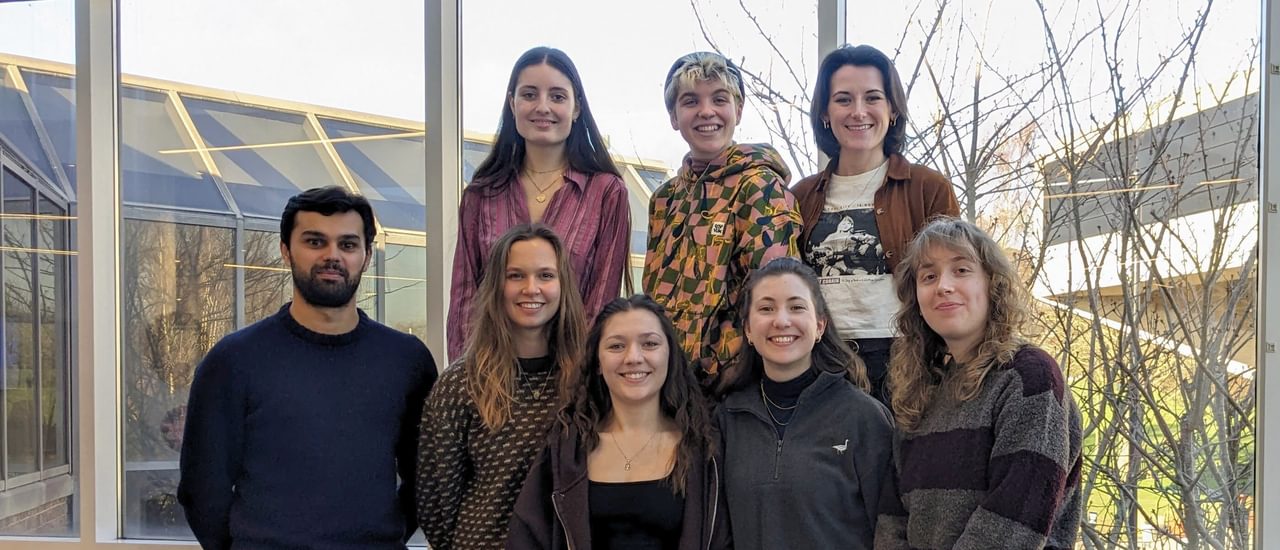 A group of eight people, all dresse4d casually, stand smiling at the camera. Behind them through the floor-length windows is glimpsed the side of the Minerva Bar and the corner of Chichester Festival Theatre.