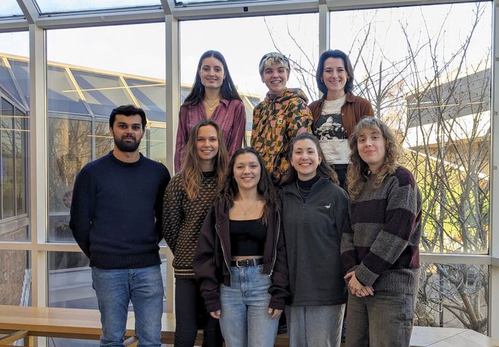 A group of eight people, all dresse4d casually, stand smiling at the camera. Behind them through the floor-length windows is glimpsed the side of the Minerva Bar and the corner of Chichester Festival Theatre.