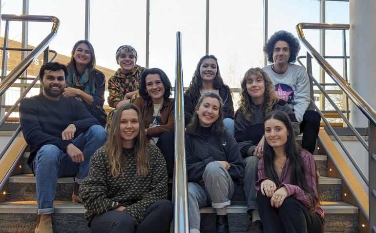A group of 10 people dressed casually and smiling sit on a flight of stairs, with a metal bannister running up between them, with an expanse of windows and sky behind them.