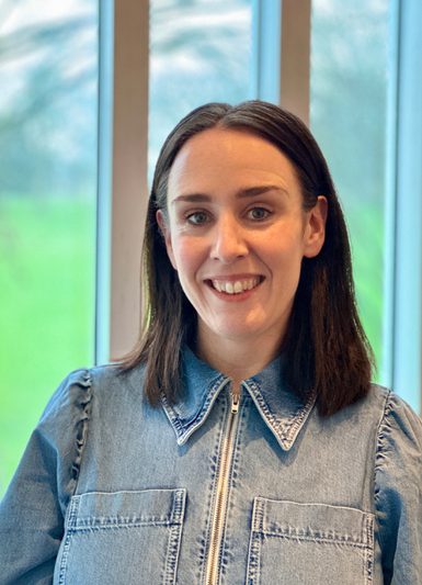 A woman with shoulder-length brown hair smiles at the camera. She wears light blue denim. Behind her is a three-panelled window through which can be glimpsed an expanse of grass and trees.
