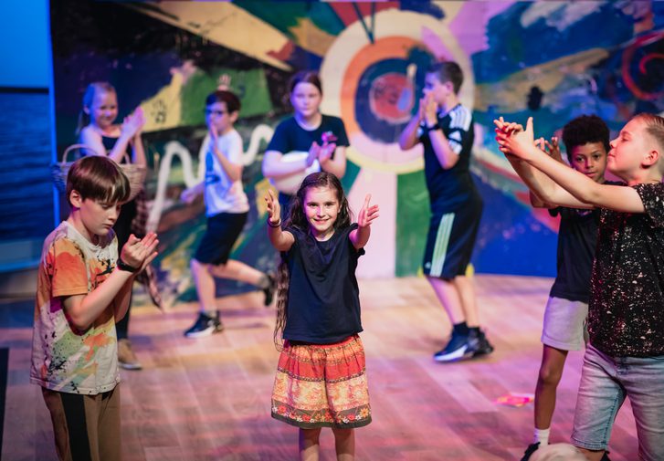 A group of young people of varying ages performing on the Minerva Theatre stage against a colourful backdrop they have painted themselves.