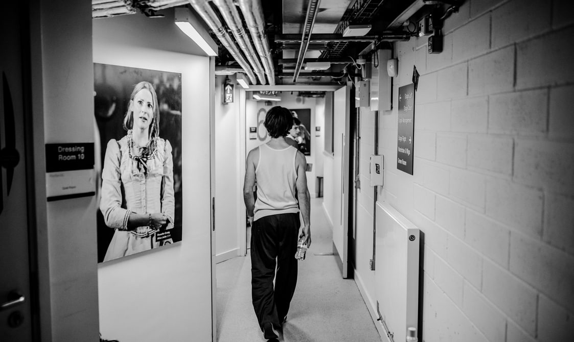 A man walks down a backstage corridor. The exposed-brick corridor is lined with pictures of actors on stage. He carries a bottle of water in his hand, his back is turned to the camera.