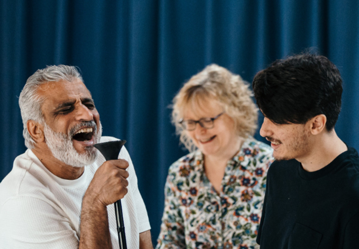 Three people taking part in a creative workshop. The man on the left is singing passionately and mimics holding a microphone on a stand, the lady in the centre laughs, and the young man on the right smiles and mimes playing a guitar.