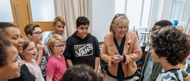 Dale Rooks dressed in a peach jacket is surrounded by children in a rehearsal space.