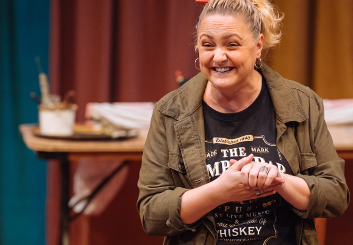 A woman with her blonde hair in a ponytail with an orange pencil tucked through it is smiling excitedly. Her hands are clasped in front of her. She is wearing a casual khaki jacket over a black graphic t-shirt. In the background, there is a table with art supplies in front of swathes of colourful curtains.