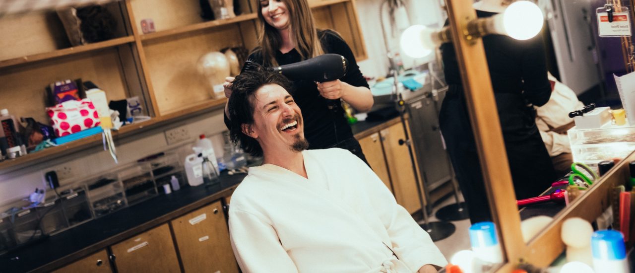 Tom Rosenthal sat at a dressing room table having his hair blow dried - he is laughing and wearing a white robe.
