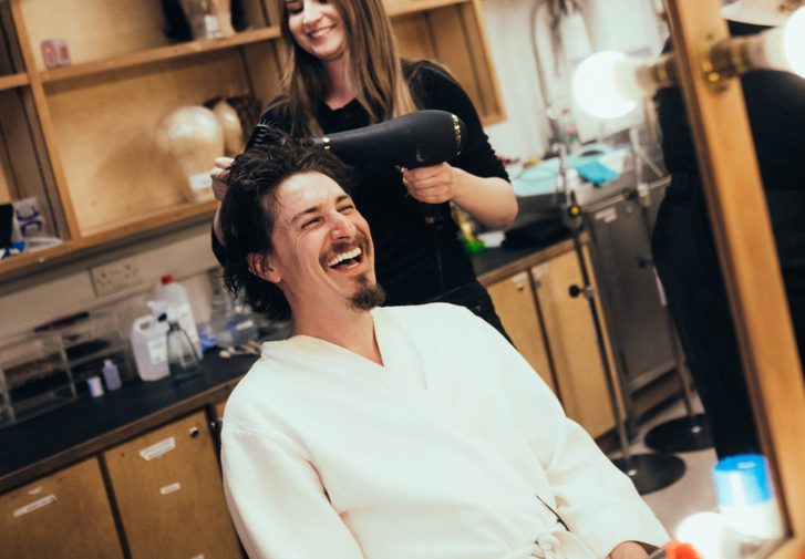Tom Rosenthal sat at a dressing room table having his hair blow dried - he is laughing and wearing a white robe.