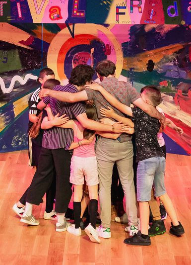 A group of children and adults are in a tight circular hug together. Arms interweave. A bright and abstract painting is behind them.
