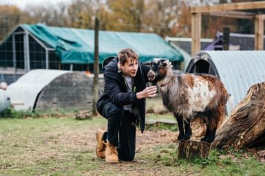 A young person crouches on the grass at a farm, petting a goat. They look engaged and happy.