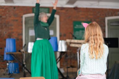 A lady wearing green performs with her arms stretched up into the air. In the foreground, a young girl listens.