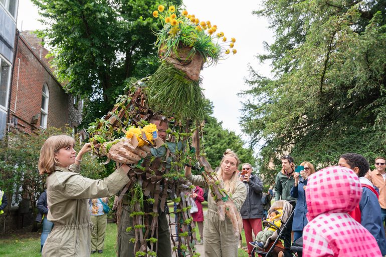 A huge puppet, operated by three puppeteers, stands outside. A curious crowd stands around watching. The puppet is woven from natural materials, with wooden arms and legs, a green, grassy beard and dandelion flowers for hair. He holds a bunch of woollen dandelions in his hand.