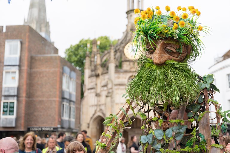 A large puppet stands outside, with a clock tower building in the background, looming above a crowd. The puppet has a woven, wooden body, green, grassy beard and dandelion flowers for hair.