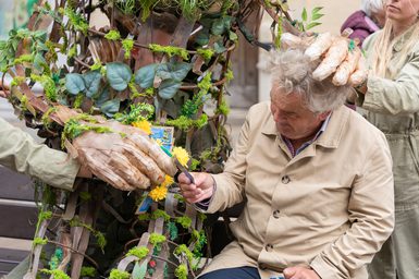 An older man sits next to a large puppet: we see the wooden, woven body of the puppet intertwined with leaves and greenery. The puppet rests one hand affectionately on the man's head and hands him a woollen dandelion flower with his oversized, wooden hand.