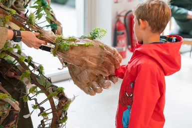 A young boy reaches out and holds hands with a large puppet. We see the puppet's woven, wooden body, intertwined with leaves and greenery, and two oversized, wooden hands. It is a tender, touching moment.