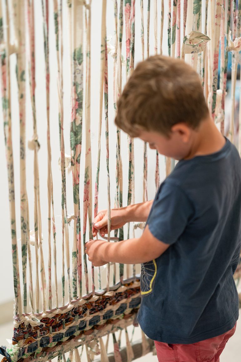 A young boy carefully adds to a large woven artwork.