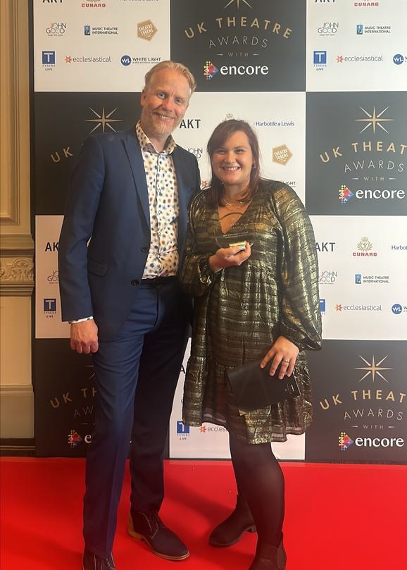 Two members of staff dressed in smart clothing stand on a red carpet in front of a UK Theatre Awards backdrop, holding a glass trophy. They look very happy and proud.