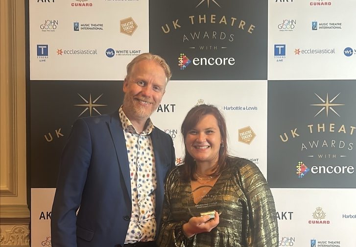 Two members of staff dressed in smart clothing stand on a red carpet in front of a UK Theatre Awards backdrop, holding a glass trophy. They look very happy and proud.