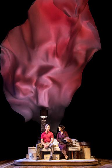Two performers are seated on a wooden bed in the centre of the stage, with a large red and purple backdrop, like a piece of fabric floating against a black background, dominating the image. The female performer on the right wears a purple suit and looks kindly at the man next to her, who wears a red polo shirt and beige trousers and looks back at her, smiling.