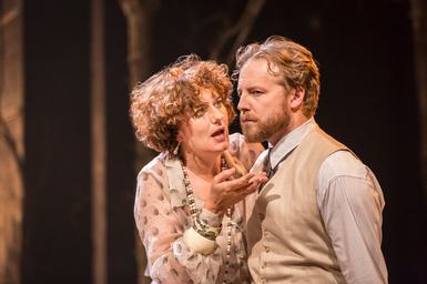 Two performers on stage. The woman on the left stands up against the man on the right. She is looking at him intently, talking, and gesturing with one hand. The man is not looking at her, instead staring off into the distance with his hands behind his back and a stern expression.