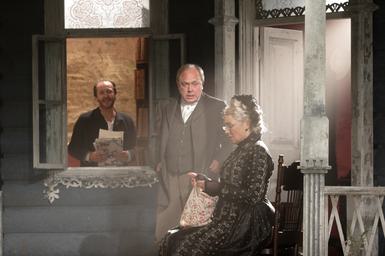 Three performers on stage. They all wear black and white and the set follows the same colour scheme, so the image almost looks black and white. A woman sits side on in the front, wearing a long-sleeved black lace dress and matching headband. She looks down at a floral bag on her lap. A man stands over her, wearing a suit, looking worried. A man can be seen through the window behind them, smiling and looking much more relaxed than the other two, holding a pile of papers.