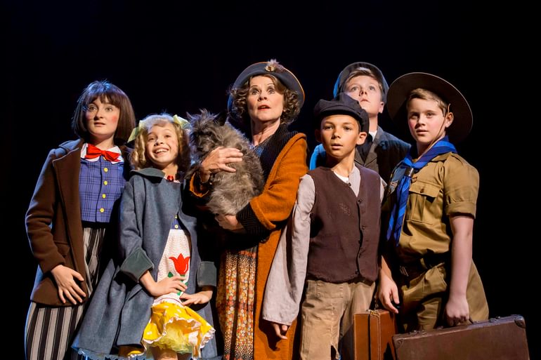 A group of children and a woman on stage. They are all looking up and out into the distance, amazed and mesmerised. They wear brightly coloured clothing. The woman stands in the centre, clutching a cat to her chest.