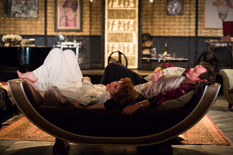 Two performers on stage. They both lie leisurely on a curved seat. They look close and in love.