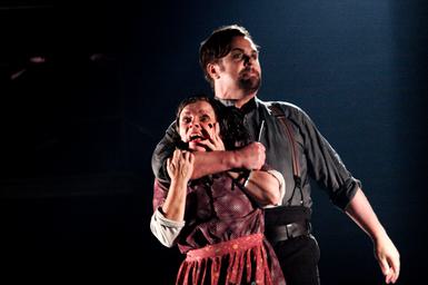 Two performers on stage. The male performer has the woman in a headlock from behind and holds a knife in his fist. He looks off to the distance with a stern expression. The woman is grasping his arm, trying to free herself, with an alarmed facial expression, holding her hand up to her face.