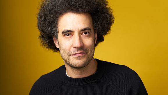 A man in a dark sweatshirt smiling in front of a yellow background