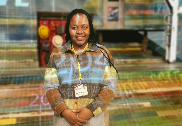 A young woman wearing a checked shirt and a CFT lanyard around her neck stands in the middle of the colourful set of The Famous Five
