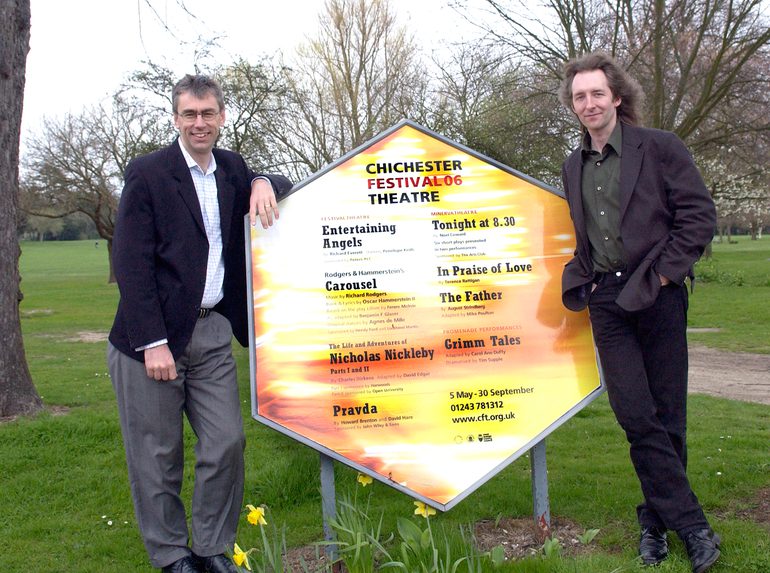 Alan Finch on the left, and Jonathan Church on the right, stand either side of an octagonal display board bearing the titles of the Festival 2016 shows, in Oaklands Park.
